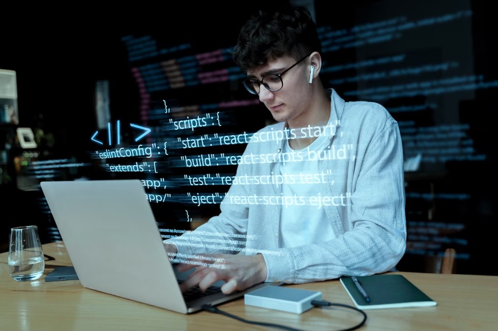 a young male in glasses and earbuds using a laptop to work with codes